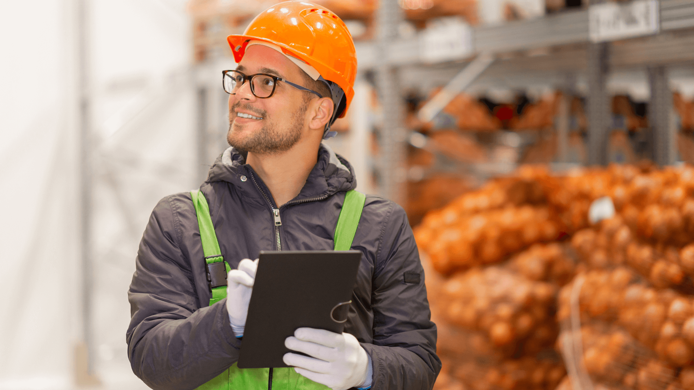 A smiling industrial worker wearing an orange hard hat and safety vest, holding a tablet and looking upwards in a warehouse setting, symbolizing efficient uniform program management and industrial uniform solutions.