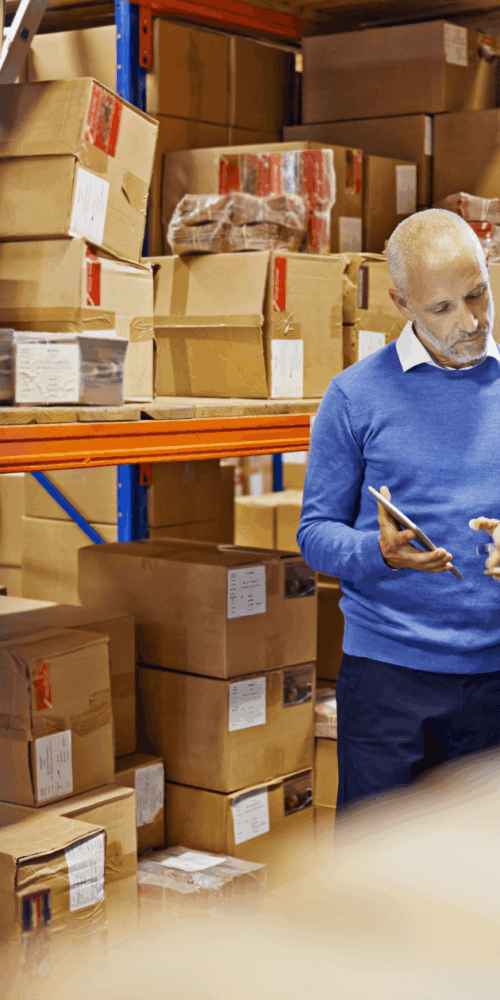 Two professionals, a man and a woman, are discussing information on a tablet within a large warehouse filled with stacked boxes, illustrating strategic uniform procurement and uniform program management.