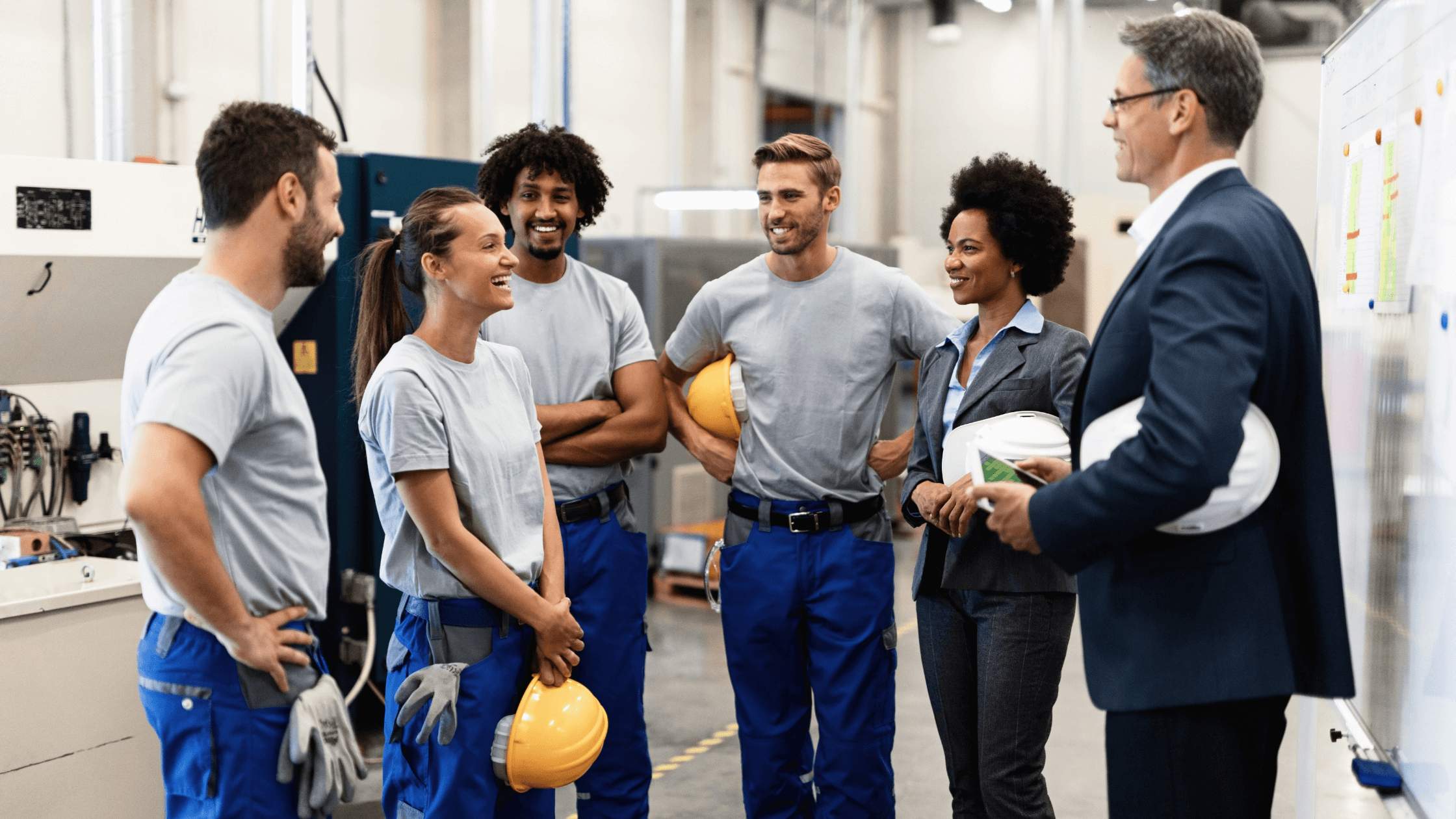 A group of diverse employees in branded uniforms and safety gear, including hard hats, smiling and talking to a supervisor in a professional setting, demonstrating positive team interaction and a successful managed uniform program.