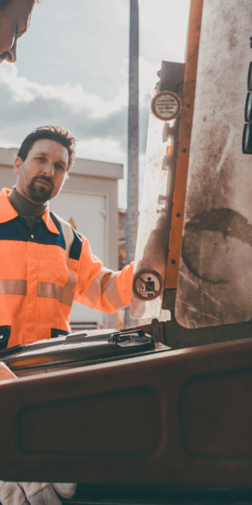 Two public sector workers in bright orange high-visibility uniforms with reflective stripes standing next to a sanitation or maintenance truck, demonstrating a team equipped with professional and safe uniforms.