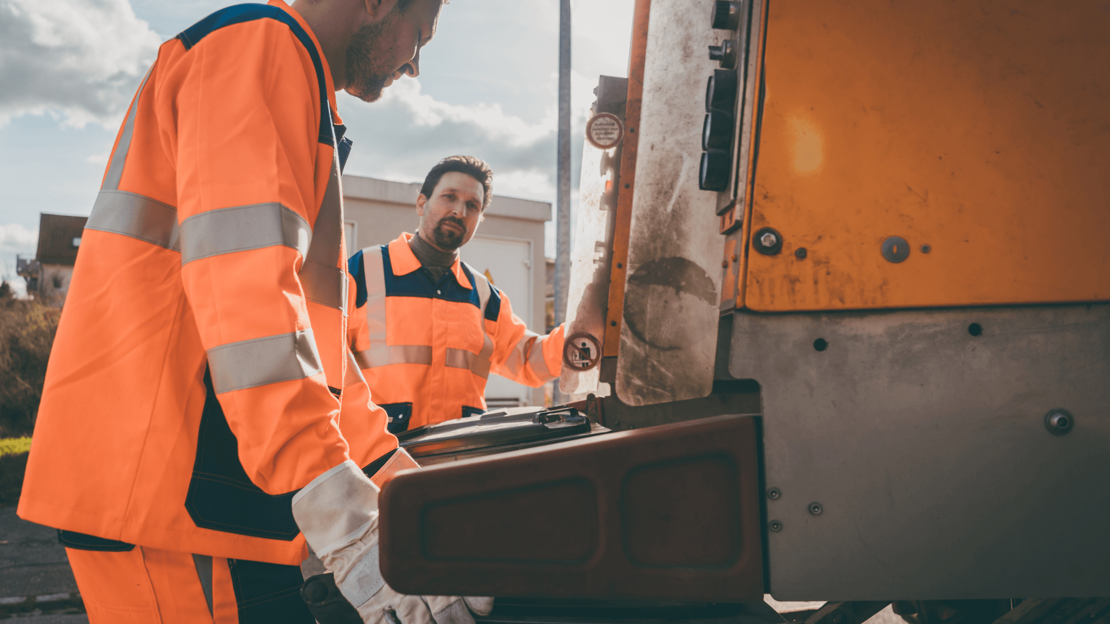 Two public sector workers in bright orange high-visibility uniforms with reflective stripes standing next to a sanitation or maintenance truck, demonstrating a team equipped with professional and safe uniforms.