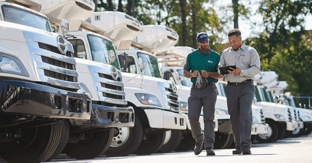 Two uniformed procurement or operations managers review data on a tablet while standing in front of a fleet of trucks, representing the strategic control and TCO analysis enabled by modern uniform management software for logistics and distribution teams.