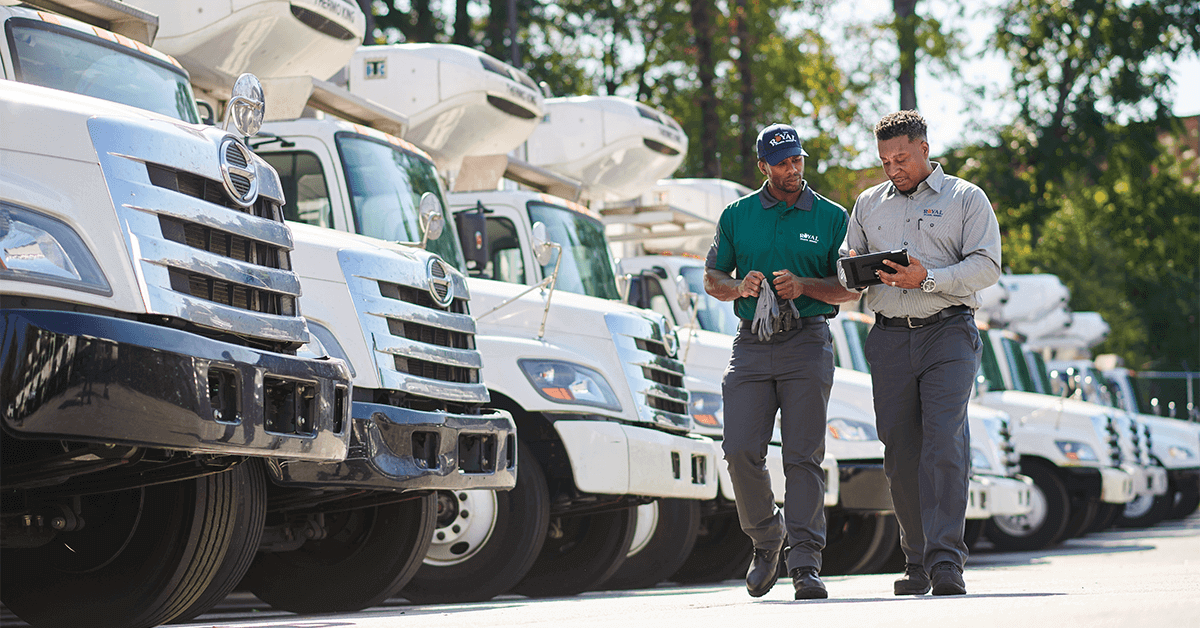 Two uniformed procurement or operations managers review data on a tablet while standing in front of a fleet of trucks, representing the strategic control and TCO analysis enabled by modern uniform management software for logistics and distribution teams.