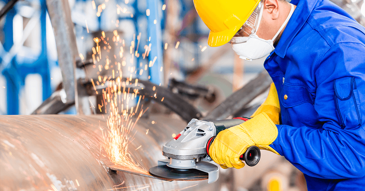 Industrial worker wearing blue fire-resistant (FR) coveralls, a yellow hard hat, safety glasses, and yellow leather gloves while grinding metal, demonstrating the need for verified NFPA 2112 or ASTM F1506-compliant apparel to protect against flash fire and sparks.