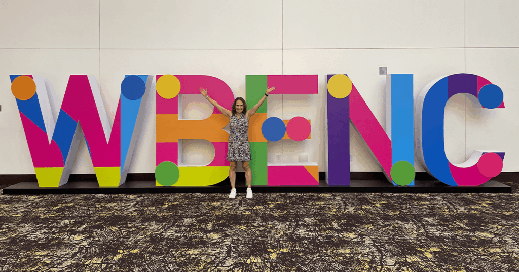A certified woman business owner stands with arms raised in front of large, colorful letters spelling "WBENC" (Women's Business Enterprise National Council), representing the benefits and national importance of WBE certification for supplier diversity in procurement.