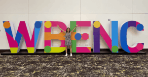 A certified woman business owner stands with arms raised in front of large, colorful letters spelling "WBENC" (Women's Business Enterprise National Council), representing the benefits and national importance of WBE certification for supplier diversity in procurement.