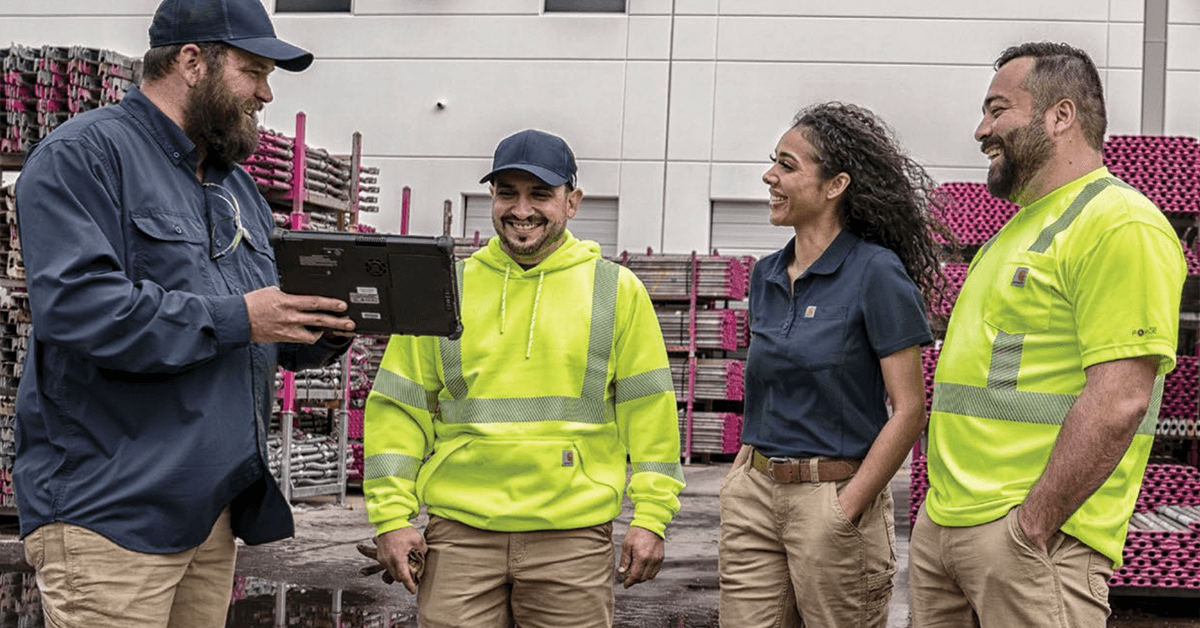 Diverse team of four industrial workers in professional uniforms (long-sleeve, polo, Hi-Vis) reviewing digital information on a tablet in a distribution setting, symbolizing the use of uniform management software for TCO analysis, compliance, and team communication in large B2B operations.