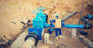 Utility worker in high-visibility safety vest and water-resistant uniform adjusting large blue water pipes in a dug-out construction site, illustrating the need for waterproof and durable apparel in water/wastewater operations.