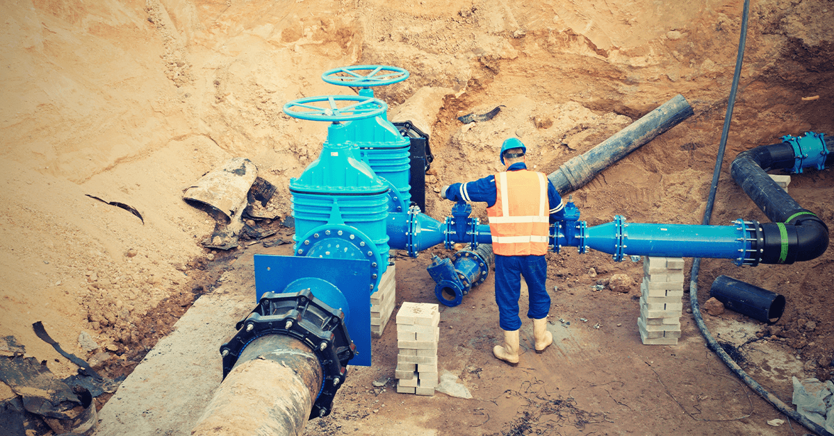 Utility worker in high-visibility safety vest and water-resistant uniform adjusting large blue water pipes in a dug-out construction site, illustrating the need for waterproof and durable apparel in water/wastewater operations.